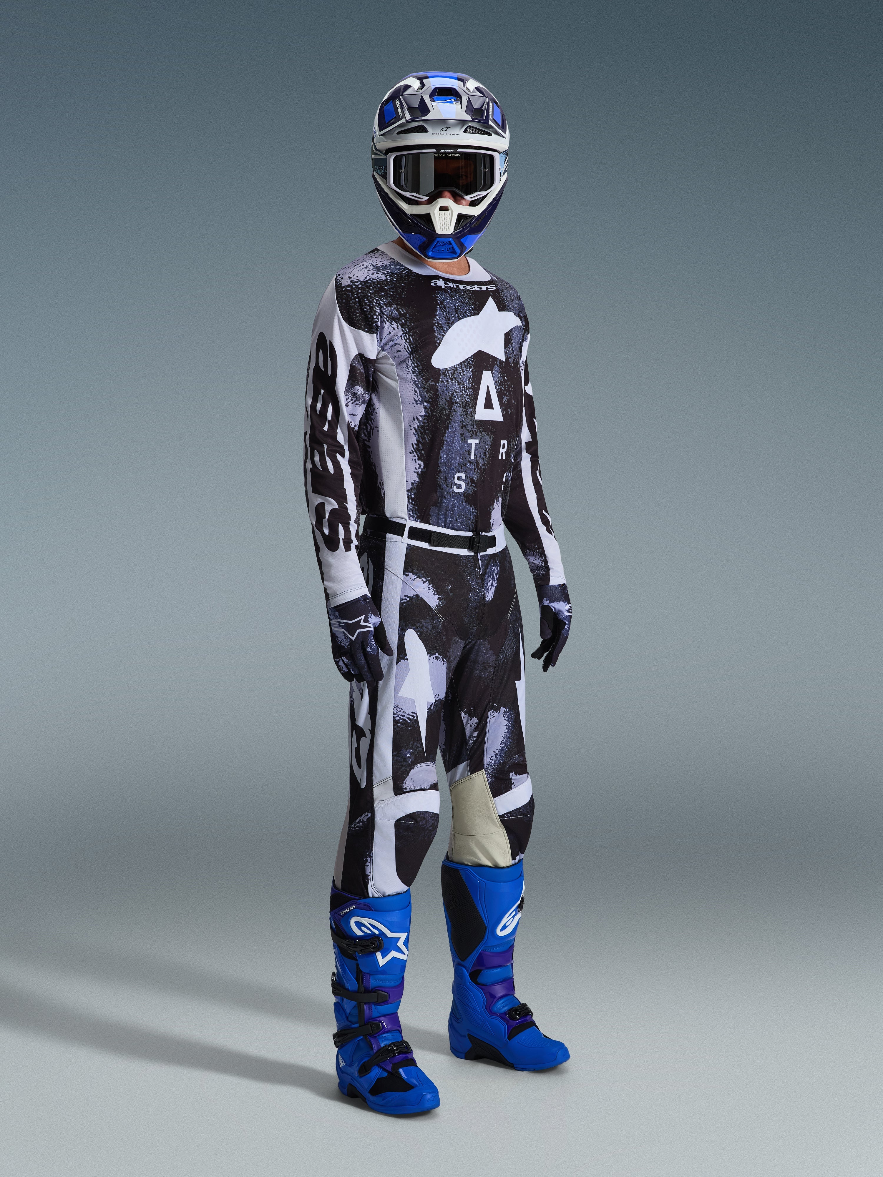 Motocrossförare som bär Alpinestars 2026 Racer Lahnd-tröja och matchande byxor i Iron Camo, med ett svartvitt grafiskt mönster, kombinerat med blå stövlar, en blå och vit hjälm och svarta handskar, komplett racingutrustning för terrängkörning.