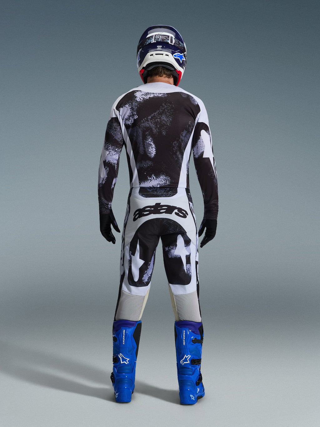 Alpinestars 2026 Racer Lahnd byxor, motocrossbyxor, Iron Camo grått och svart mönster, vy bakifrån av förare i komplett racingutrustning med matchande tröja och blå stövlar, designade för terrängprestanda.