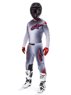 Motocross rider wearing full Alpinestars 2025 Supertech Lipan racing gear set, Gray and Orange Fluo, gray jersey with large orange chest logo, matching pants with orange accents, black helmet, red gloves, and white boots, professional off-road racing outfit
