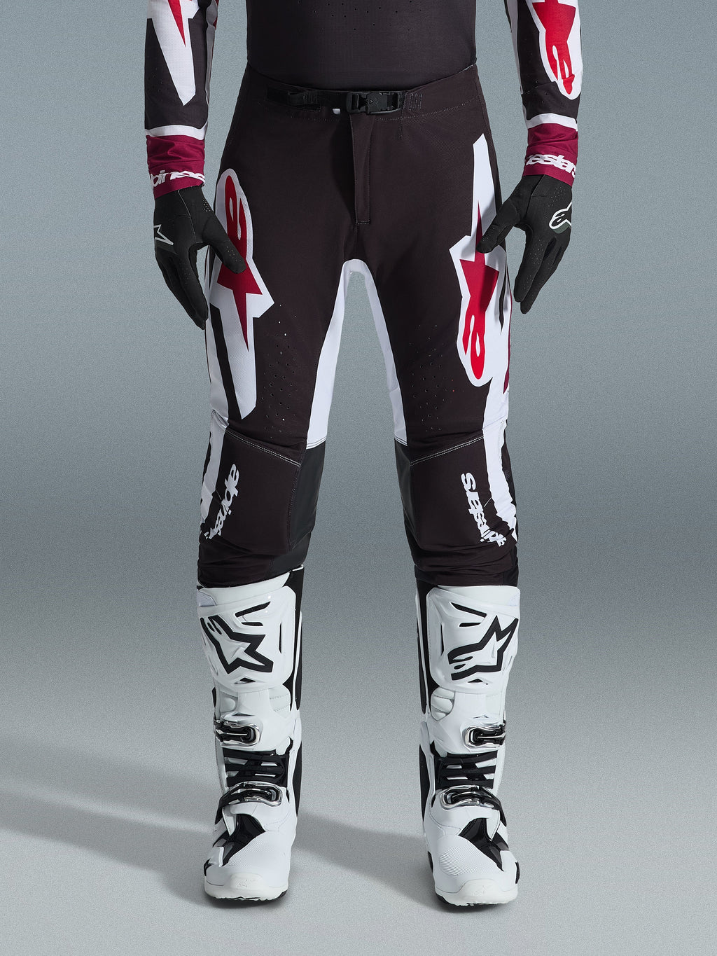 Alpinestars 2026 Supertech AFD Vista byxor, motocrossbyxor, svarta, röda och vita, med stora grafiska logotyper på låren och knäna, bärs med vita racingstövlar och svarta handskar, atletisk race-ready-design för elit-offroadtävlingar