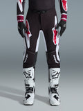 Alpinestars 2026 Supertech AFD Vista byxor, motocrossbyxor, svarta, röda och vita, med stora grafiska logotyper på låren och knäna, bärs med vita racingstövlar och svarta handskar, atletisk race-ready-design för elit-offroadtävlingar