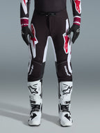 Alpinestars 2026 Supertech AFD Vista byxor, motocrossbyxor, svarta, röda och vita, med stora grafiska logotyper på låren och knäna, bärs med vita racingstövlar och svarta handskar, atletisk race-ready-design för elit-offroadtävlingar