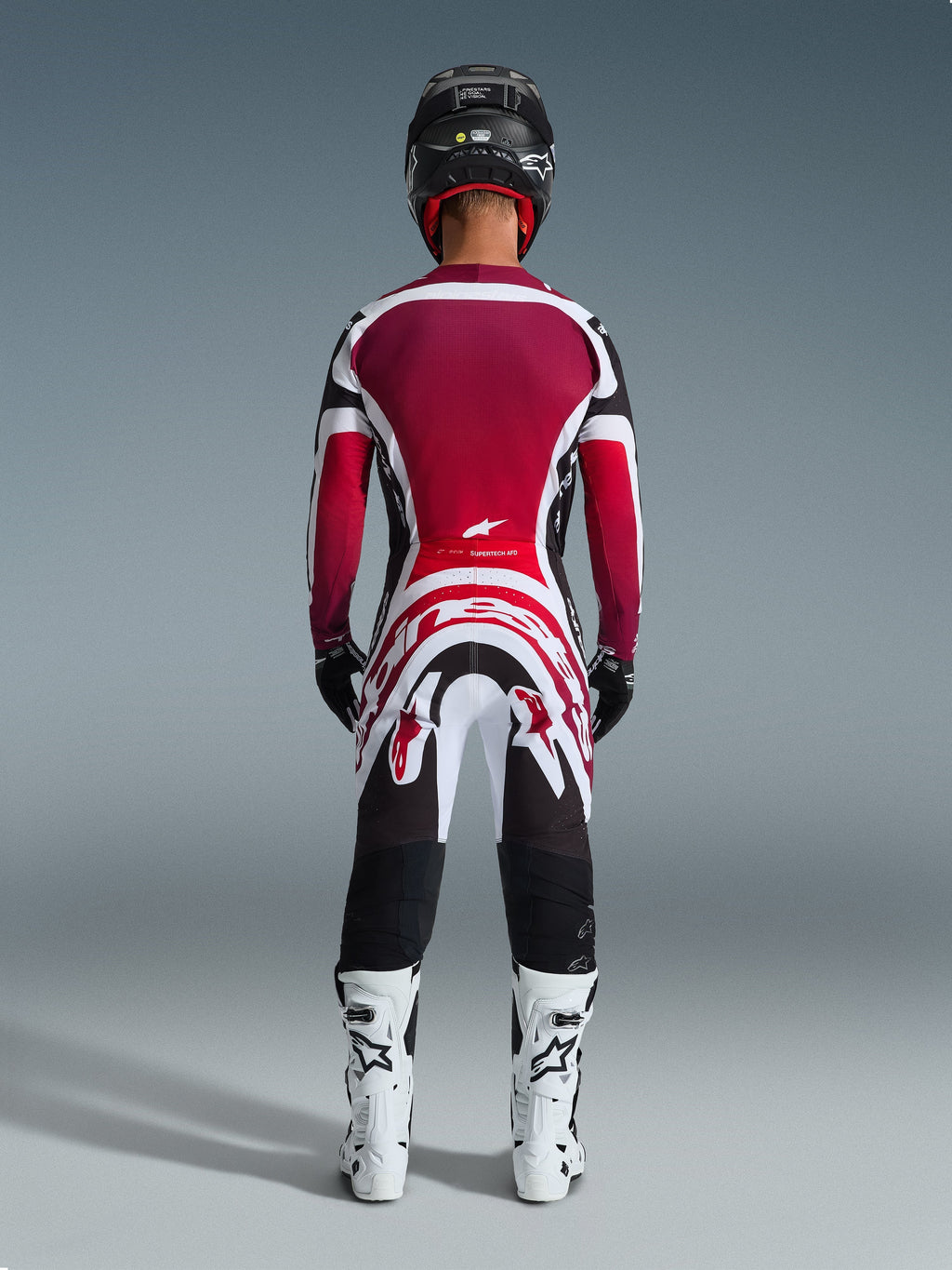 Motocrossförare sedd bakifrån iklädd Alpinestars 2026 Supertech AFD Vista-byxor, svart, röd och vit, matchande tröja, vita racingstövlar och svart hjälm, komplett professionell utrustning för terrängkörning