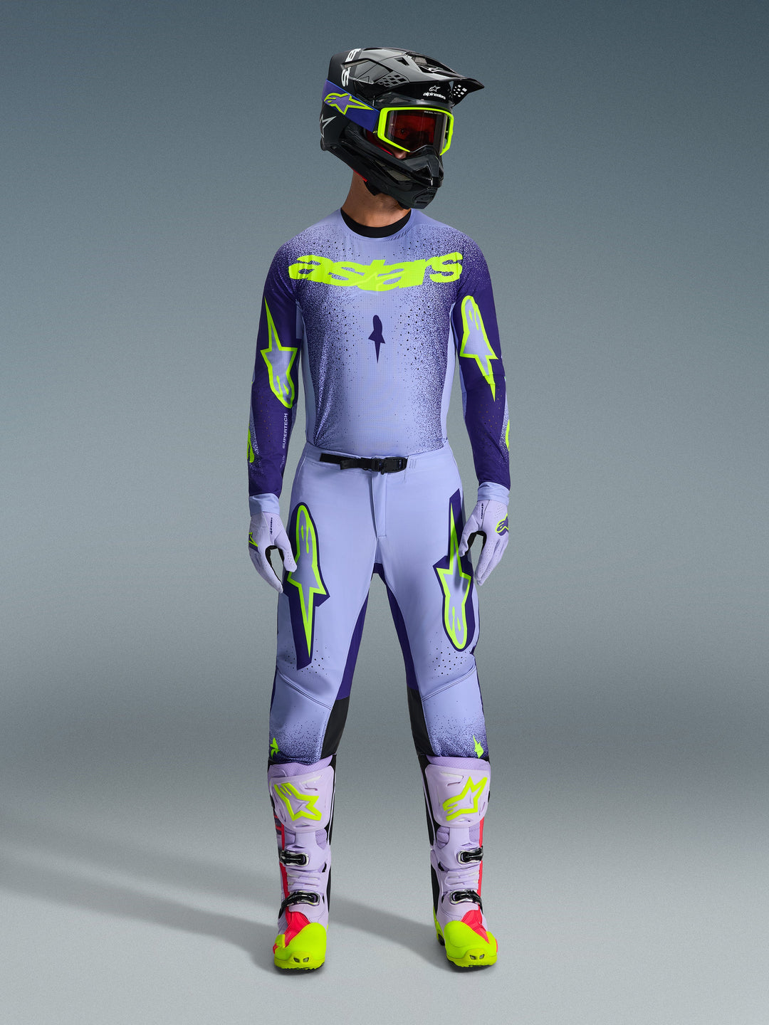 Alpinestars 2026 Supertech Scenz utrustningsset, motocross-outfit, ljuslila och neongul-grön, komplett skyddsutrustning inklusive svart hjälm med lila accenter, tröja med tydlig neongul logotyp, matchande byxor med stjärngrafik och koordinerade stövlar med rosa detaljer.