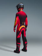 Motocrossförare sedd bakifrån klädd i Alpinestars 2026 Supertech Scenz-byxor i rött, svart och gult, matchande tröja med framträdande gul logotyp, svarta och röda motocross-stövlar, svart hjälm, komplett professionellt racingställ för terrängkörning.