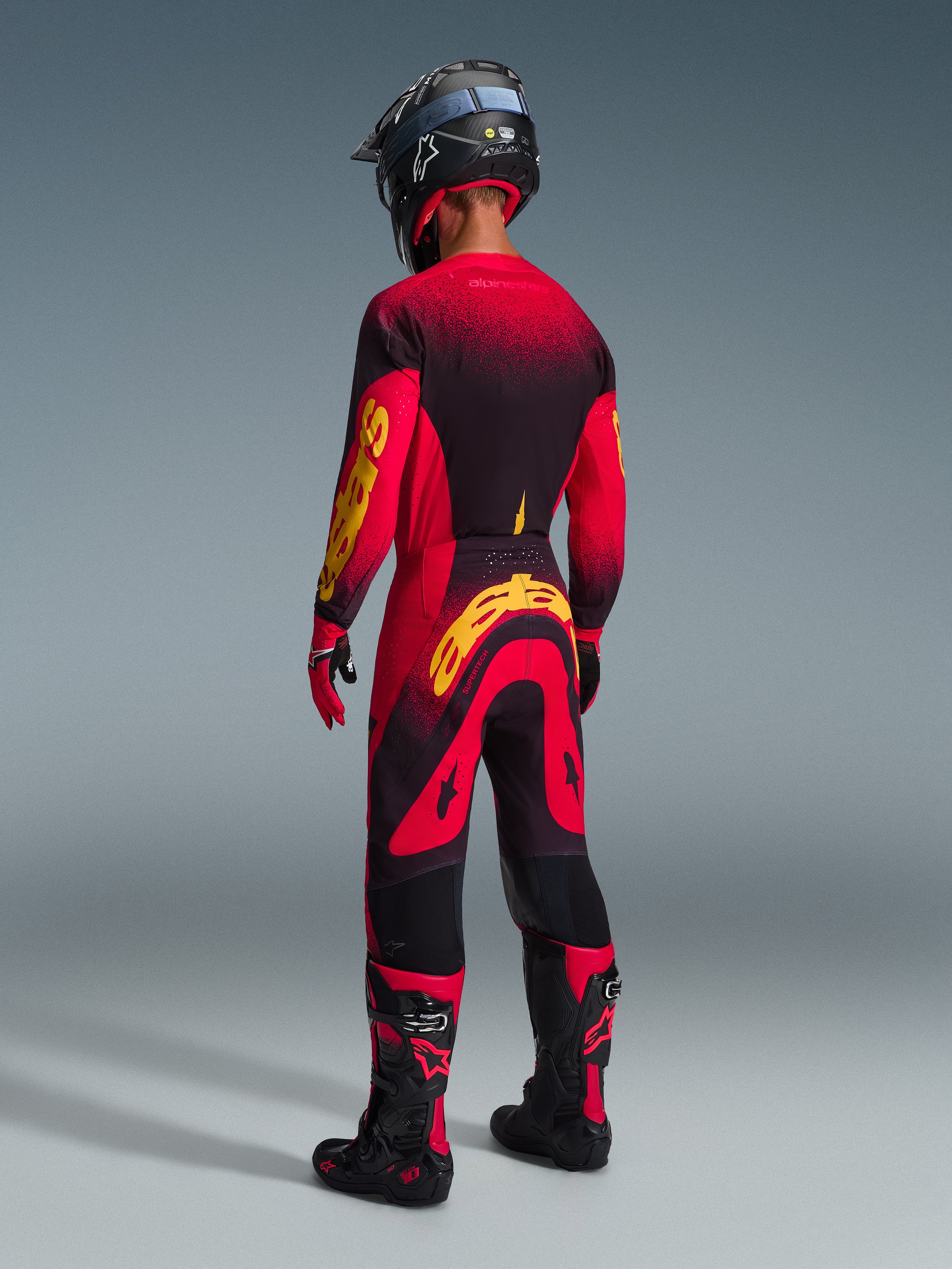 Motocrossförare sedd bakifrån klädd i Alpinestars 2026 Supertech Scenz-byxor i rött, svart och gult, matchande tröja med framträdande gul logotyp, svarta och röda motocross-stövlar, svart hjälm, komplett professionellt racingställ för terrängkörning.