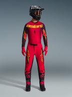 Alpinestars 2026 Supertech Scenz byxor, motocrossbyxor, röda, svarta och gula, bärs av en förare i en komplett racingutrustning, matchande röd och svart tröja med gula logotyper, svart hjälm, röda handskar och svarta stövlar, atletisk pro-fit-design för terrängtävling