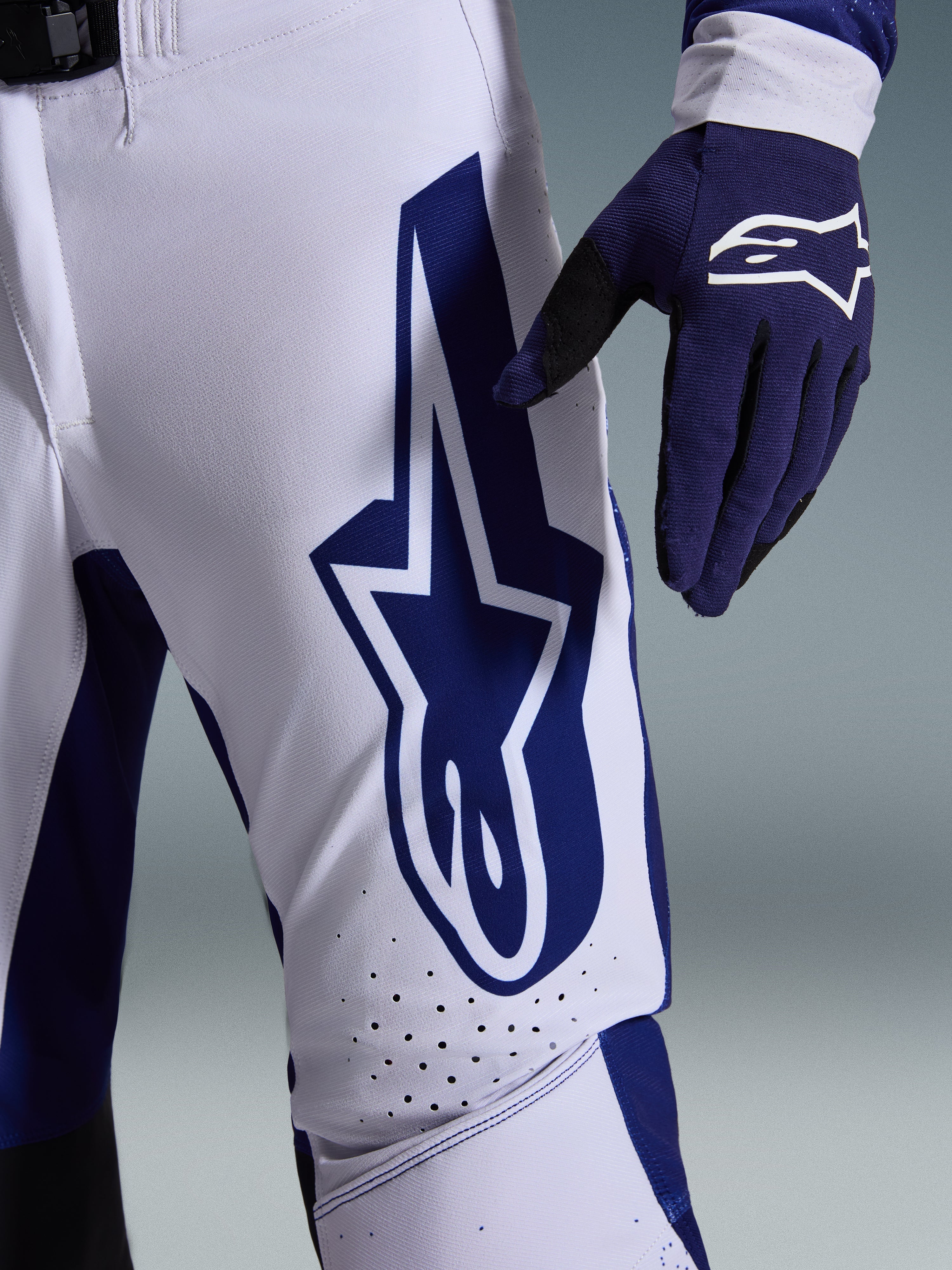 Alpinestars 2026 Supertech Scenz Pants, Motocross Pants, Light Gray and Dark Blue, close-up view of the thigh showing a large dark blue Alpinestars logo, perforated ventilation zones, and stretch rip-stop fabric, worn with a matching dark blue motocross glove
