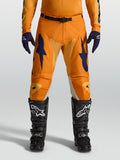 Alpinestars Racer Amber byxor, orange och blåa motocrossbyxor för herrar med racingpassform, slitstarkt 600D-polytyg, visas på modell med svarta motorcykelstövlar.