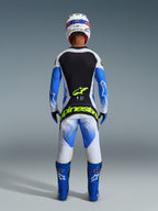 Motocrossförare sedd bakifrån i komplett racingutrustning, 2026 Techstar Knif-byxor i UCLA Blue, svart och neongult, matchande långärmad tröja med svarta och vita paneler, blå off-road-stövlar, vit hjälm, med framträdande neongul Alpinestars-logotyp på sätet.