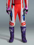 Alpinestars 2026 Techstar Knif-byxor, motocrossbyxor i Orange Fluo och ljusgrått med svarta detaljer, visas med lila motocross-stövlar och handskar, med blå stjärnlogotyper och flerpanelkonstruktion för terrängprestanda.