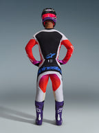 Motocrossförare sedd bakifrån i 2026 Techstar Knif-byxor och matchande tröja, ljusgrå och svart design med starka orangea och lila detaljer, blå Alpinestars-logotyp på sätet, lila stövlar och hjälm, professionellt utrustningsset för terrängkörning.