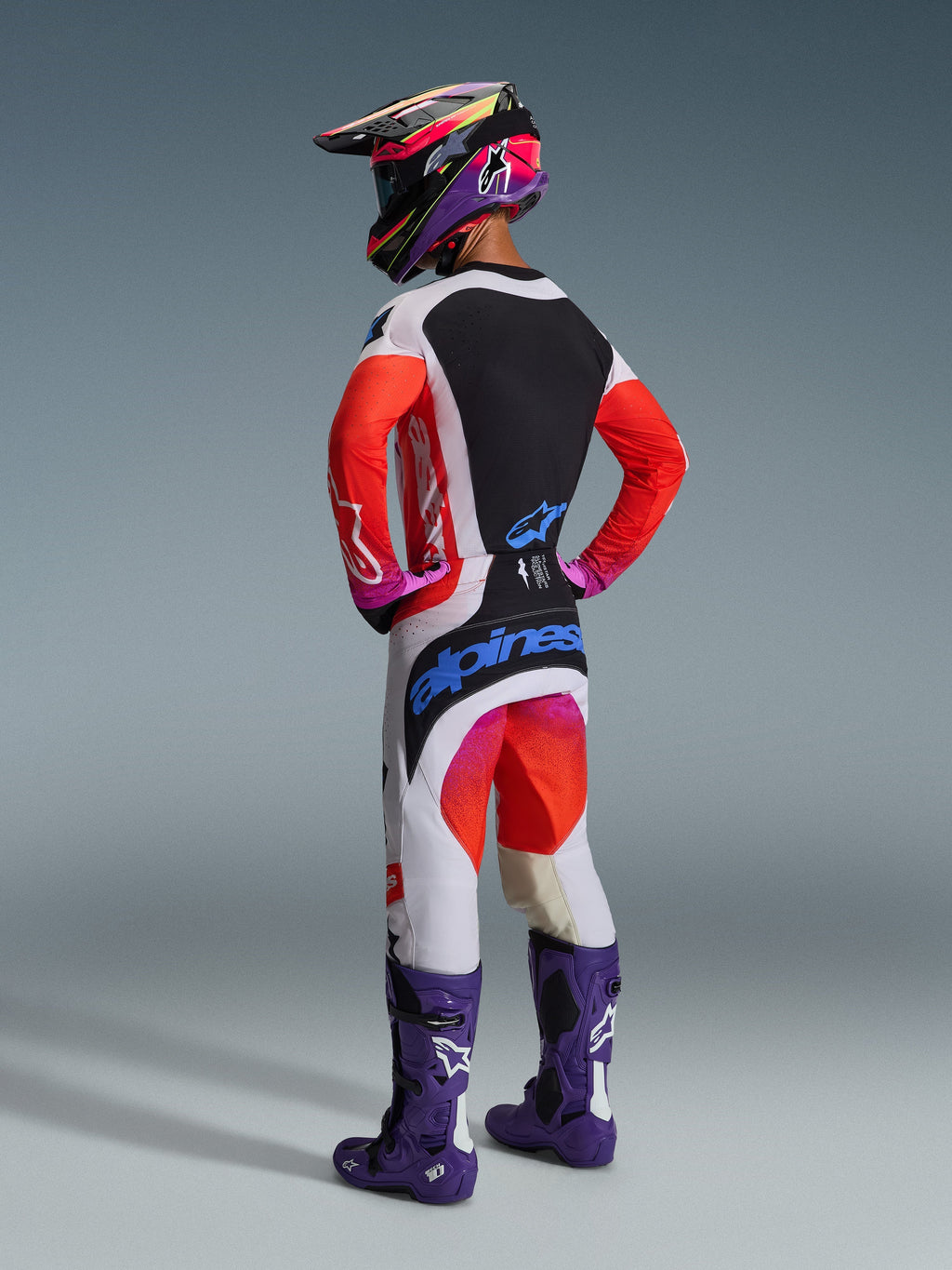 Motocrossförare sedd bakifrån i komplett racingutrustning, Alpinestars 2026 Techstar Knif-byxor i ljusgrått och orange fluo med blå logotyp, matchande tröja med orangea ärmar, lila Tech 10-stövlar och en färgglad racinghjälm, koordinerad professionell off-road-outfit