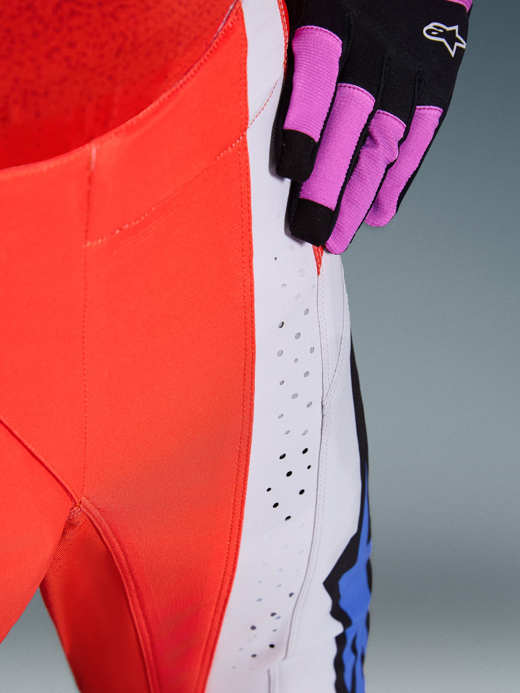 Alpinestars 2026 Techstar Knif-byxor, motocrossbyxor, fluoriserande orange och ljusgrå med blå detaljer, närbild som visar laserskurna ventilationsperforeringar på låret, bärs med en svart och lila motocrosshandske