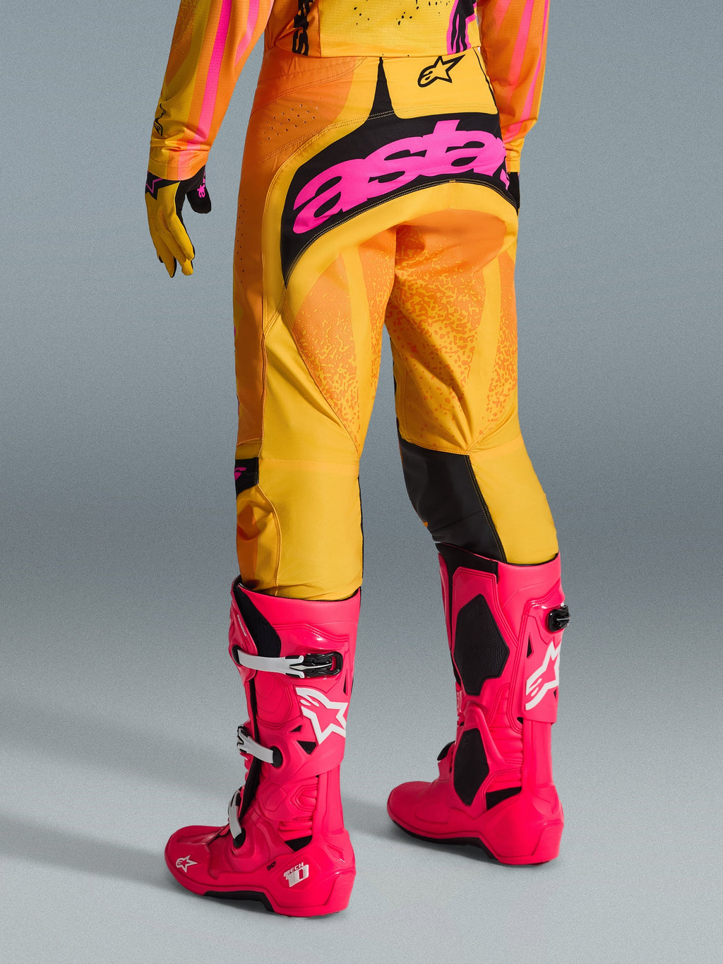 Alpinestars 2026 Techstar Nomur-byxor, motocrossbyxor, orange, gula och rosa, vy bakifrån, med stor rosa "astars"-logotyp på sätet, kombinerade med klarrosa Tech 10-stövlar och matchande tröja, högpresterande racingutrustning för motocross
