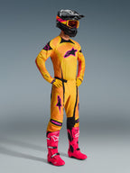 Motocrossförare iklädd Alpinestars 2026 Techstar Nomur-byxor i Orange Yellow Pink, komplett racingställ med matchande tröja och handskar, starkt rosa stövlar, svart hjälm med orange goggles, helkroppsbild mot en grå bakgrund, professionell outfit för terrängkörning