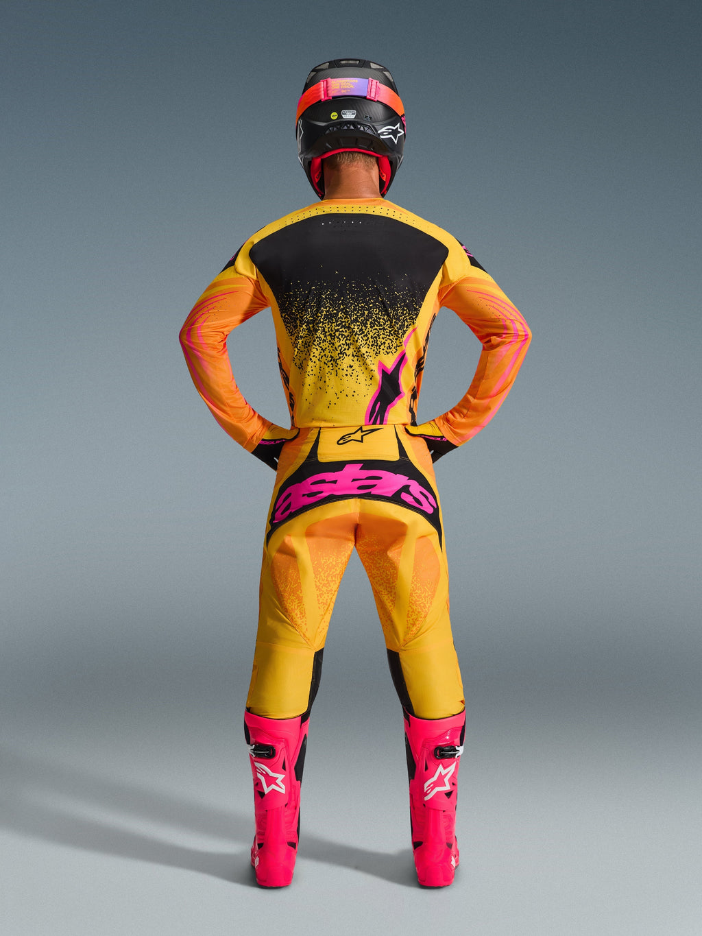 Vy bakifrån av en motocrossförare iförd Alpinestars 2026 Techstar Nomur-byxor i Orange Yellow Pink, med en stor rosa logotyp på sätet, kombinerat med en matchande gul och svart tröja, klarrosa stövlar och en svart och rosa hjälm.