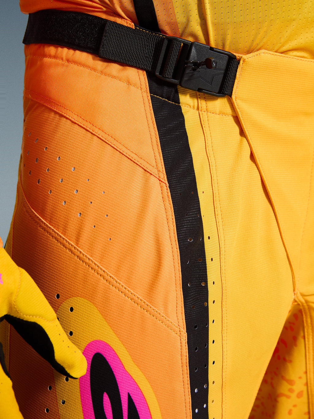 Alpinestars 2026 Techstar Nomur byxor, motocrossbyxor, orange, gul och rosa, närbild av midjan med det magnetiska SX2-spännet, svart justeringsrem och laserskurna ventilationsperforeringar på slitstarkt ripstop-material