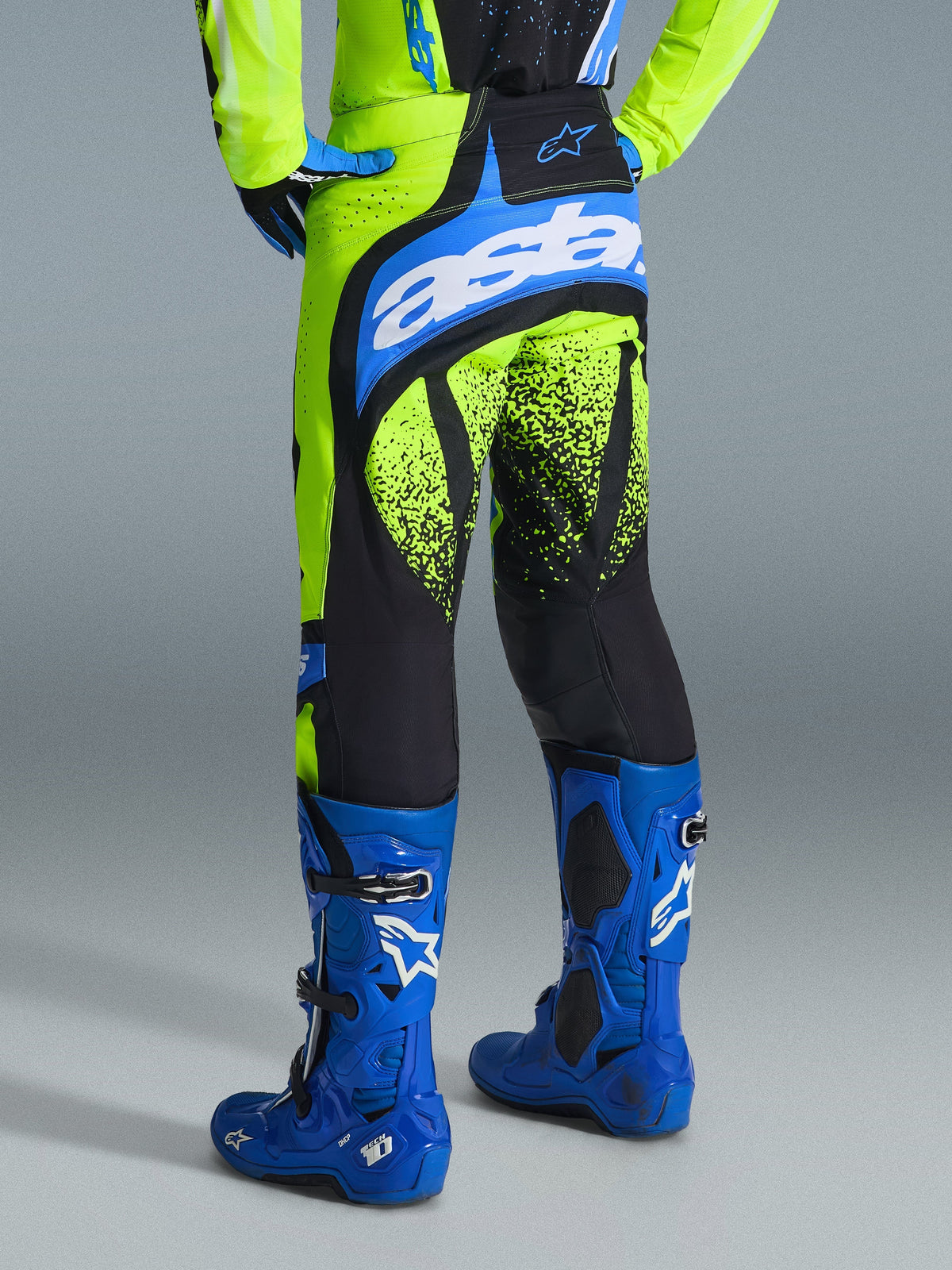Alpinestars 2026 Techstar Nomur-byxor, motocrossbyxor, gul fluo och UCLA-blå, vy bakifrån på förare i blå stövlar, med 'astars'-logotyp på sätet och svart stänkmönstrad grafik på benen