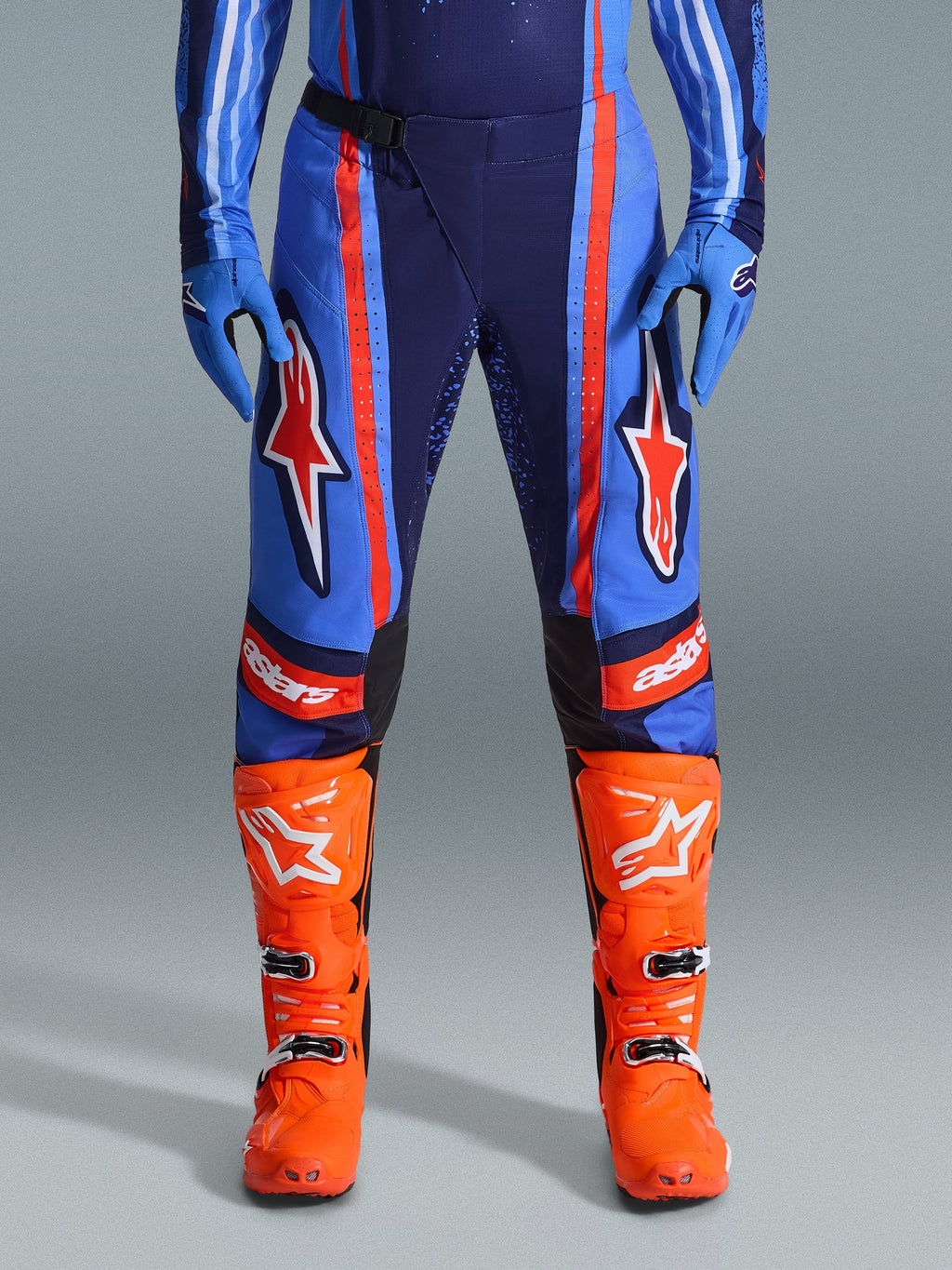 Alpinestars 2026 Techstar Nomur byxor, motocrossbyxor, mörkblå, orange och UCLA-blå, med markerade orange ränder och stora vita stjärnlogotyper på låren, bärs med klarorange racingstövlar och blå handskar, atletisk racingpassform för offroad-prestanda