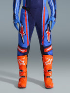 Alpinestars 2026 Techstar Nomur byxor, motocrossbyxor, mörkblå, orange och UCLA-blå, med markerade orange ränder och stora vita stjärnlogotyper på låren, bärs med klarorange racingstövlar och blå handskar, atletisk racingpassform för offroad-prestanda