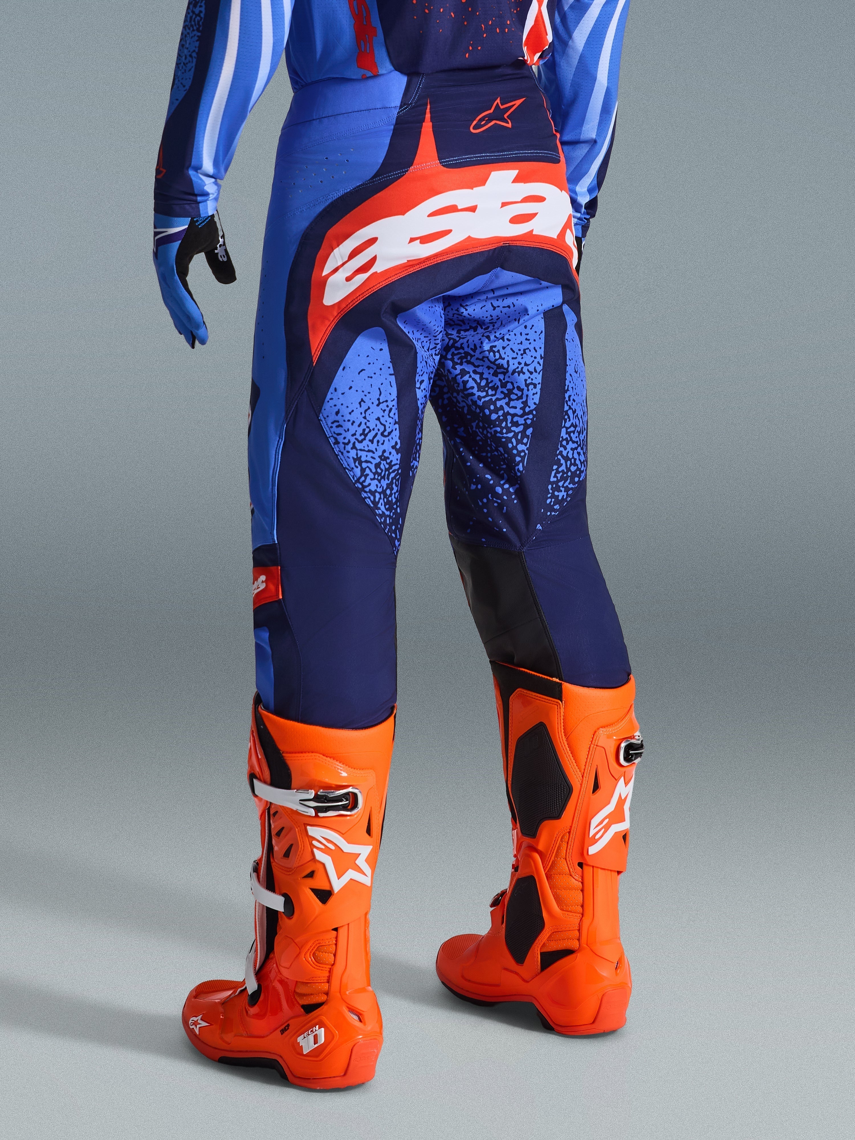 Alpinestars 2026 Techstar Nomur-byxor, motocrossbyxor, mörkblå, orange och UCLA-blå, vy bakifrån som visar vit 'Astars'-logotyp på sätet, bärs med klarorangea motocross-stövlar och matchande tröja, atletisk tävlingspassform för terrängkörning