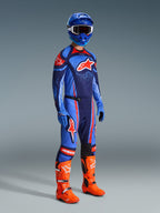 Alpinestars 2026 Techstar Nomur-byxor, motocrossbyxor, mörkblå, orange och UCLA-blå, bärs av en förare i komplett racingutrustning inklusive matchande tröja, blå hjälm och klarorangea stövlar, prestandakonstruerade för terrängkörning