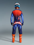 Motocrossförare sedd bakifrån bär Alpinestars 2026 Techstar Nomur-byxor i Dark Navy, orange och UCLA Blue, matchade med en tillhörande tröja och klarorangea stövlar, professionell utrustning för terrängkörning.