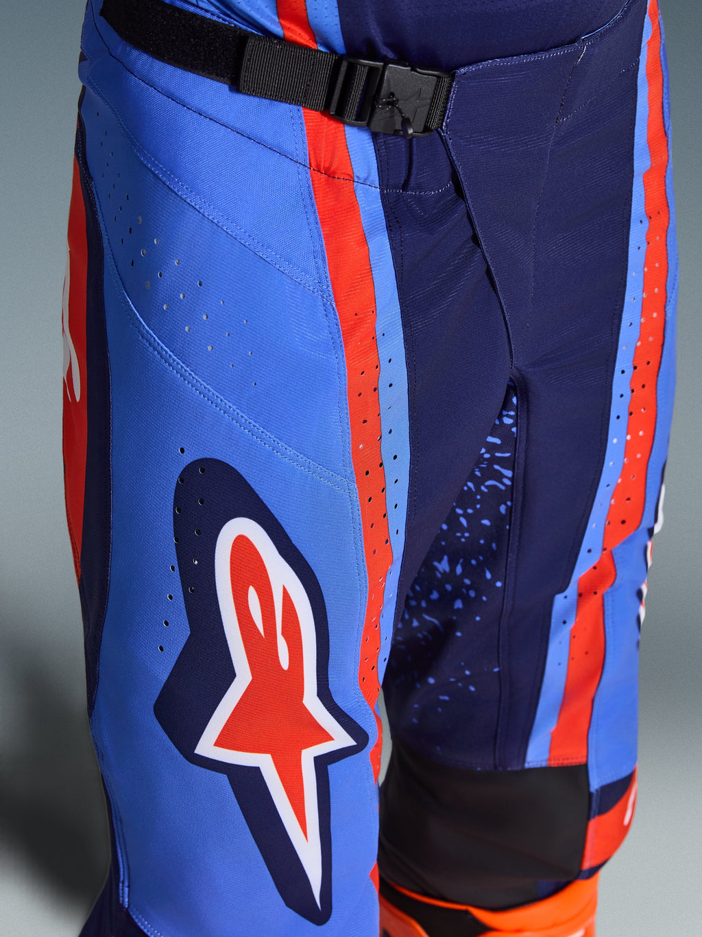 Alpinestars 2026 Techstar Nomur byxor, motocrossbyxor i mörkblått, orange och UCLA-blått, närbild som visar det magnetiska SX2-spännet i midjan, laserskurna perforeringar på låret och ikonisk stjärnlogotyp, hållbar racingutrustning för terrängkörning.