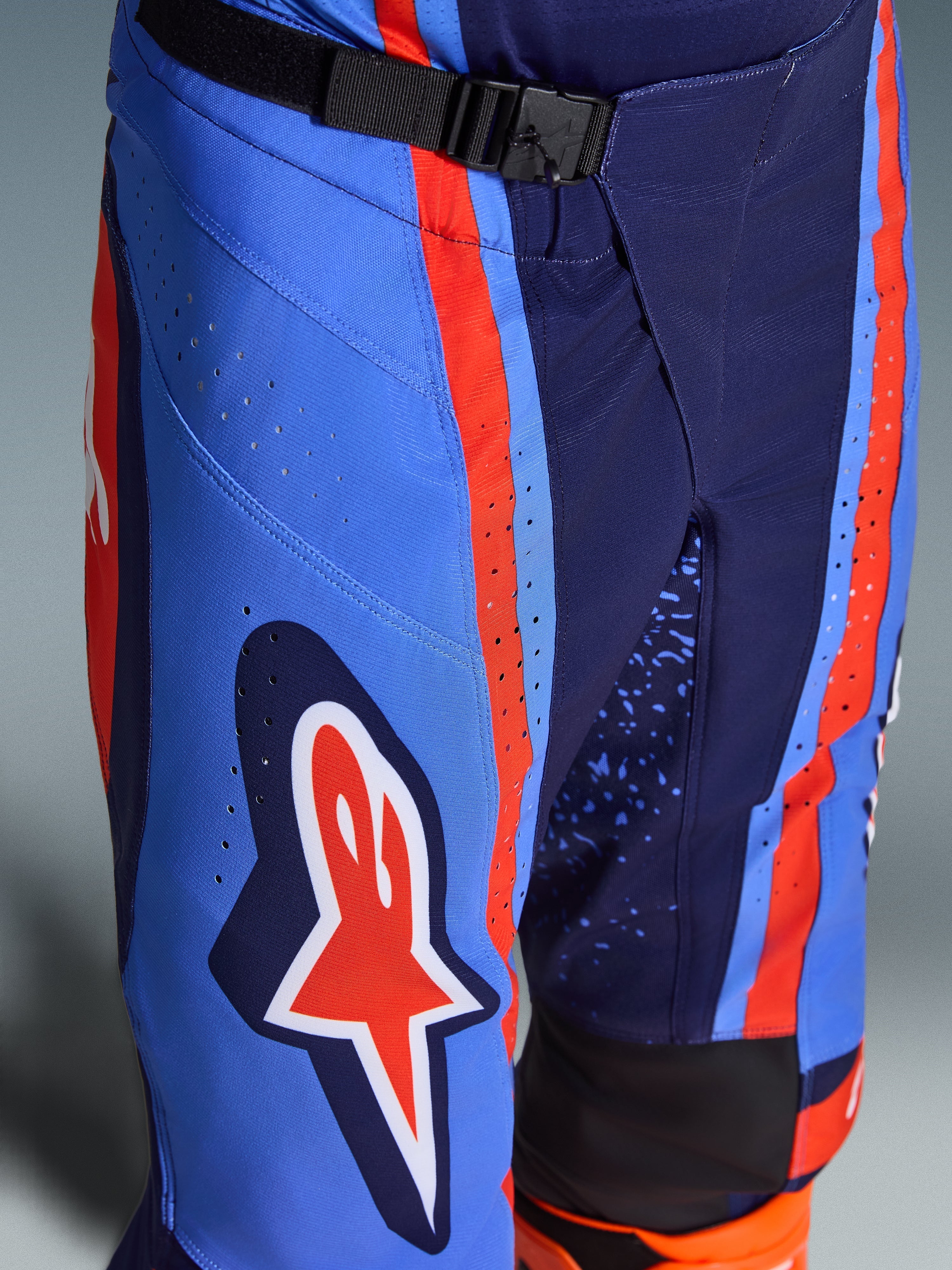 Alpinestars 2026 Techstar Nomur byxor, motocrossbyxor i mörkblått, orange och UCLA-blått, närbild som visar det magnetiska SX2-spännet i midjan, laserskurna perforeringar på låret och ikonisk stjärnlogotyp, hållbar racingutrustning för terrängkörning.