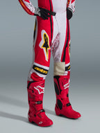 Alpinestars 2026 Techstar Nomur-byxor, motocrossbyxor, röda, vita och svarta med gula detaljer, bärs med matchande röda motocross-stövlar och svarta handskar, gul Alpinestars-logotyp och "astars"-grafik på knäna, professionell utrustning för terrängkörning.
