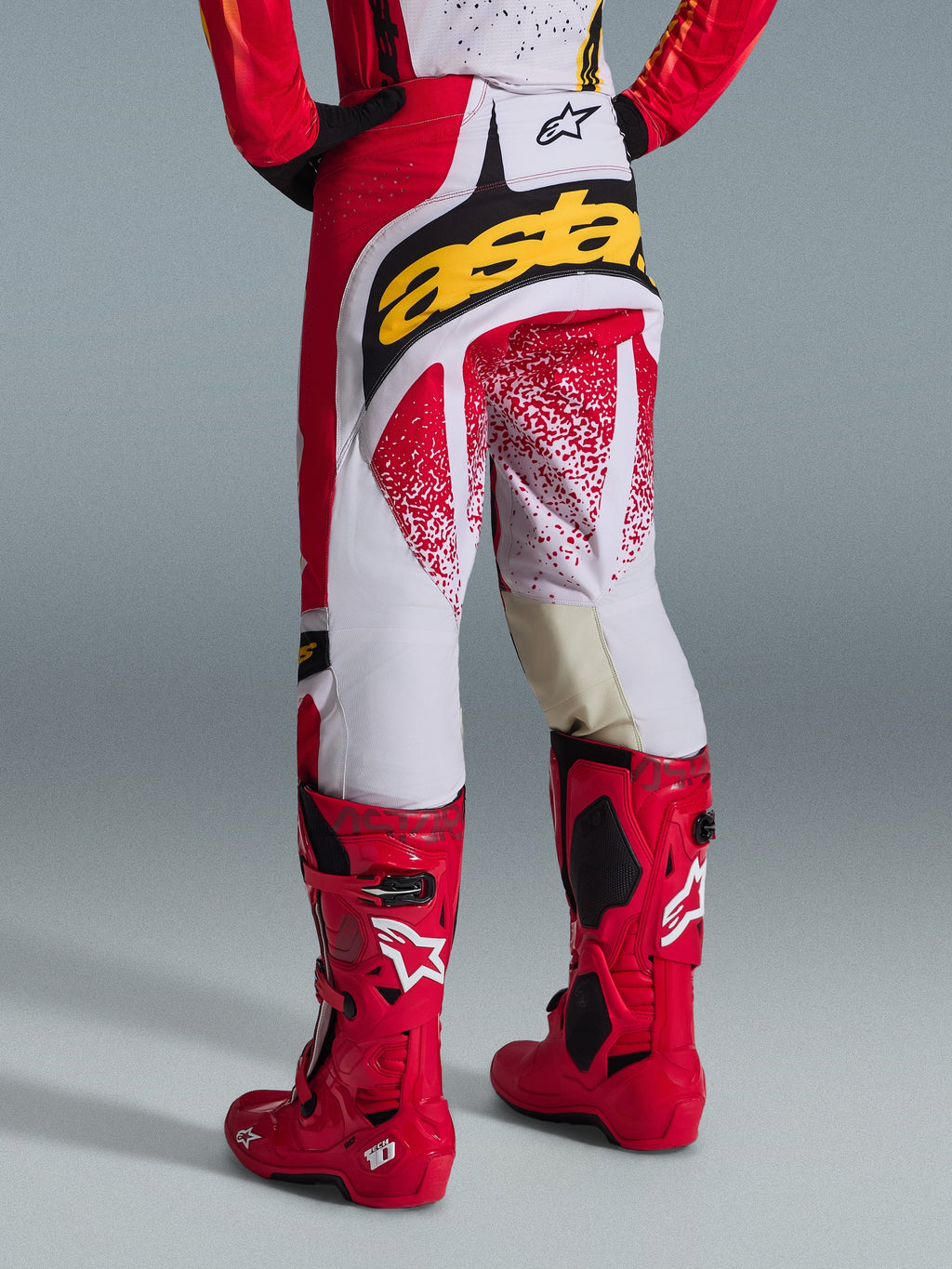 Vy bakifrån av en motocrossförare som bär Alpinestars 2026 Techstar Nomur-byxor i rött och vitt med en framträdande gul logotyp på baken, matchade med klarröda Tech 10 motocross-stövlar, studiobild mot en grå bakgrund.