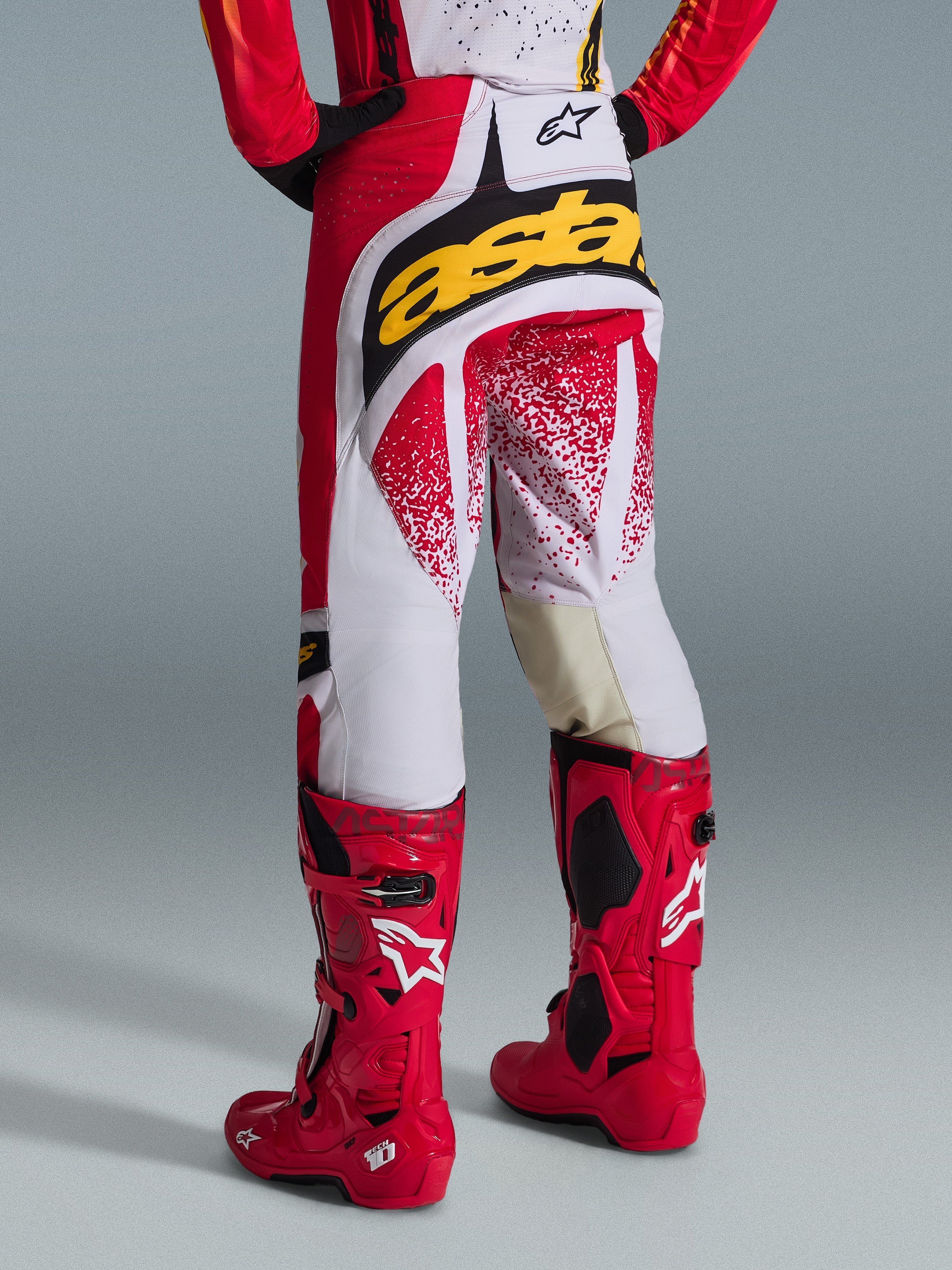 Vy bakifrån av en motocrossförare som bär Alpinestars 2026 Techstar Nomur-byxor i rött och vitt med en framträdande gul logotyp på baken, matchade med klarröda Tech 10 motocross-stövlar, studiobild mot en grå bakgrund.