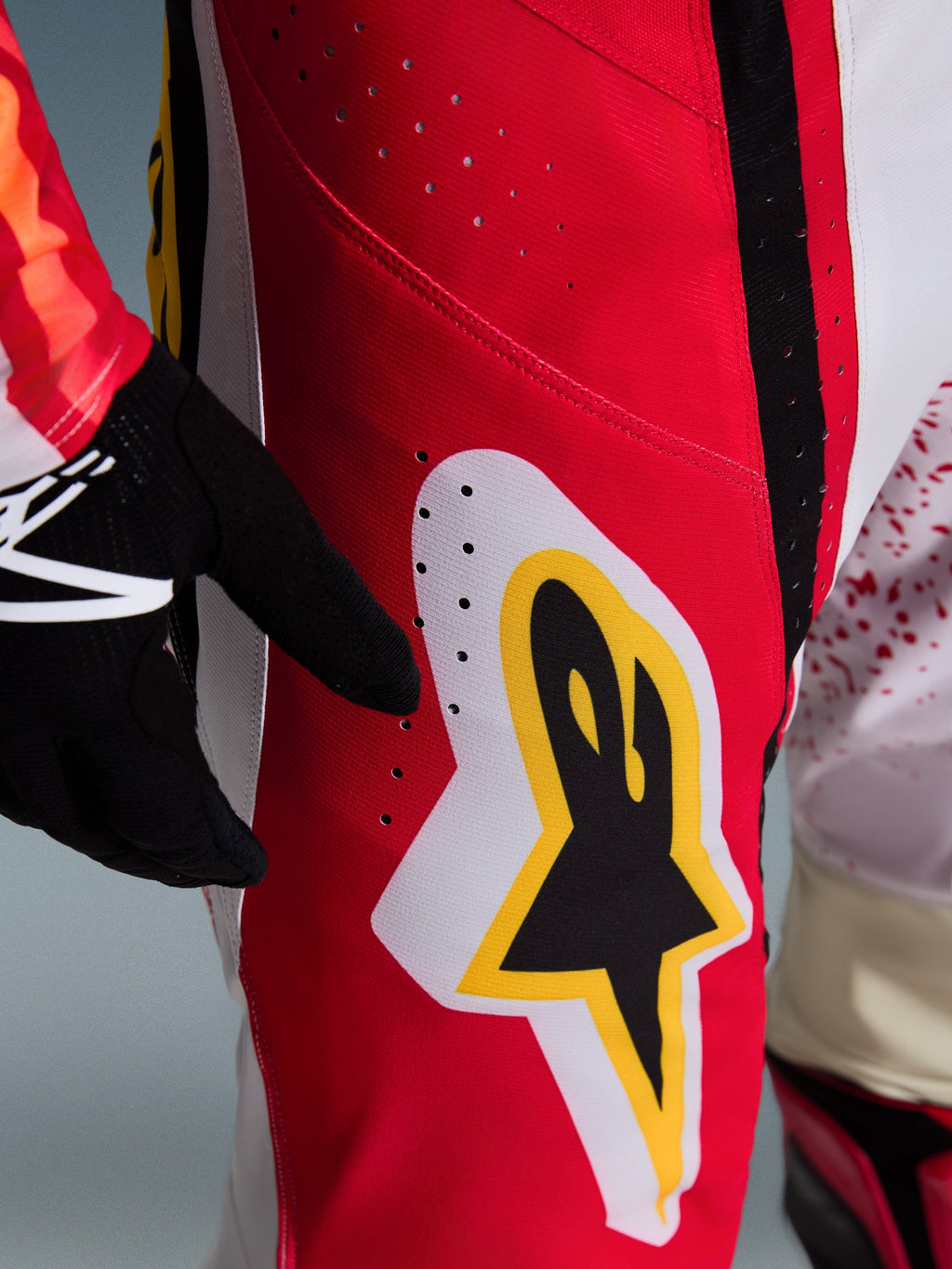 Alpinestars 2026 Techstar Nomur byxor, motocrossbyxor, röda med vita och gula detaljer, närbild på lårområdet med laserskurna ventilationshål och en stor stjärnlogotyp, matchas med svarta handskar och en färgstark tröja för terrängkörning
