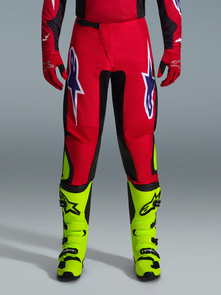 Alpinestars 2026 Fluid Grid byxor, motocrossbyxor, röda med svarta paneler och neongula detaljer, stora vita och lila stjärnlogotyper på låren, bärs med röda handskar och neongula stövlar, slitstark konstruktion för terrängkörning