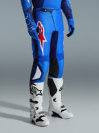 Alpinestars 2026 Fluid Grid byxor, motocrossbyxor, blå och röda, bärs med vita motocross-stövlar och blå handskar, med en stor röd stjärnlogotyp på låret och svarta paneler på knänas insida, designade för terrängkörning