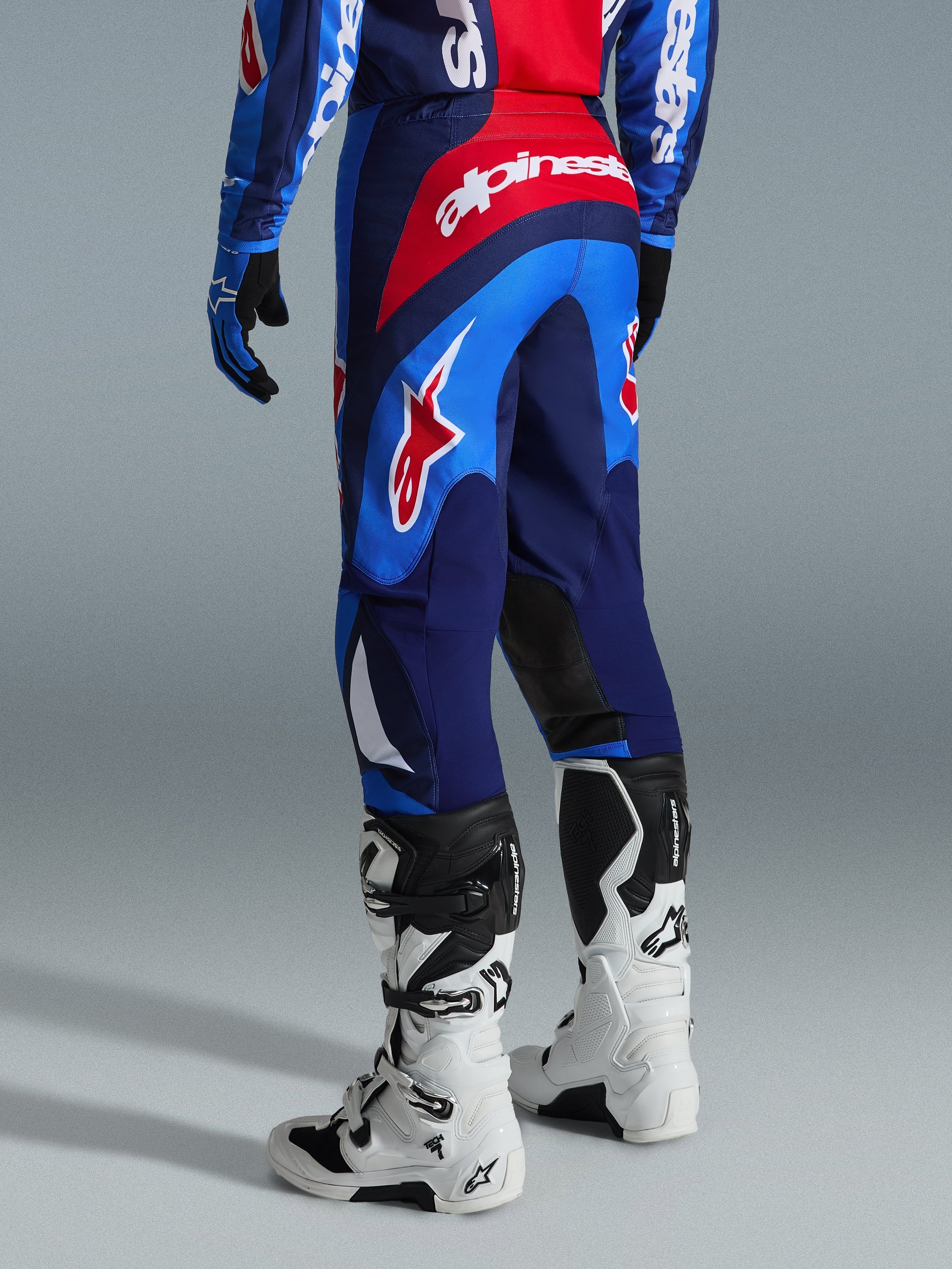 Alpinestars 2026 Fluid Grid byxor, motocrossbyxor, blå och röda, vy bakifrån som visar tydlig Alpinestars-branding och ergonomisk design, bärs med vita och svarta Tech 7-stövlar och matchande tröja, högpresterande utrustning för terrängkörning