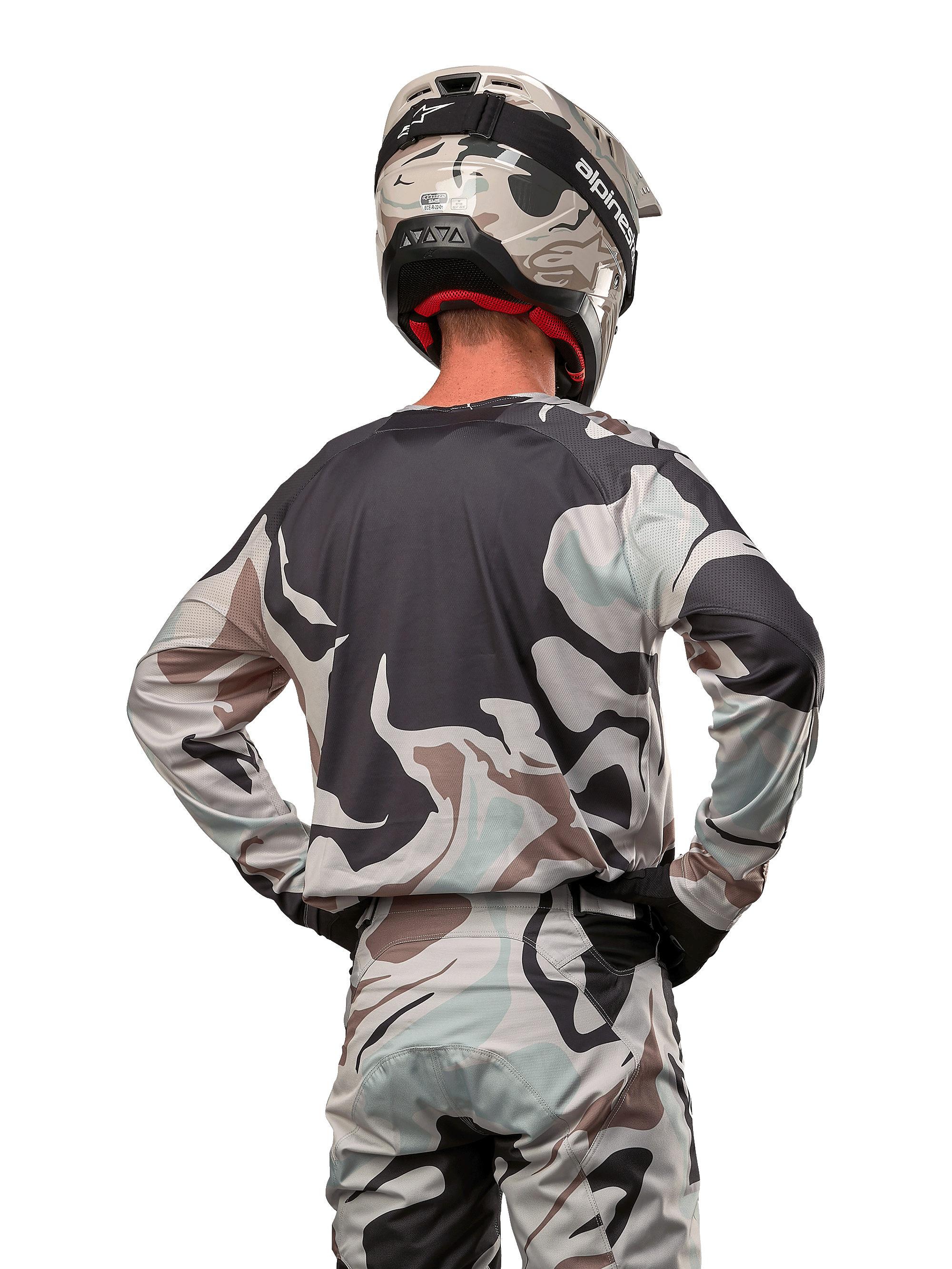 Motocrossförare sedd bakifrån, Alpinestars 2024 Racer Tactical Jersey i Iron Camo Dust Gray, matchande kamouflagebyxor och hjälm, händerna på höfterna, koordinerad racingutrustning för terrängkörning