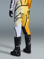 Alpinestars 2026 Fluid Apex byxor, motocrossbyxor, gul och svart färgtoning med vita och orangea detaljer, sedda bakifrån, bärs med matchande tröja och svarta Tech 7-stövlar, med en framträdande svart Alpinestars-logotyp på sätet, prestandautrustning för terrängkörning.