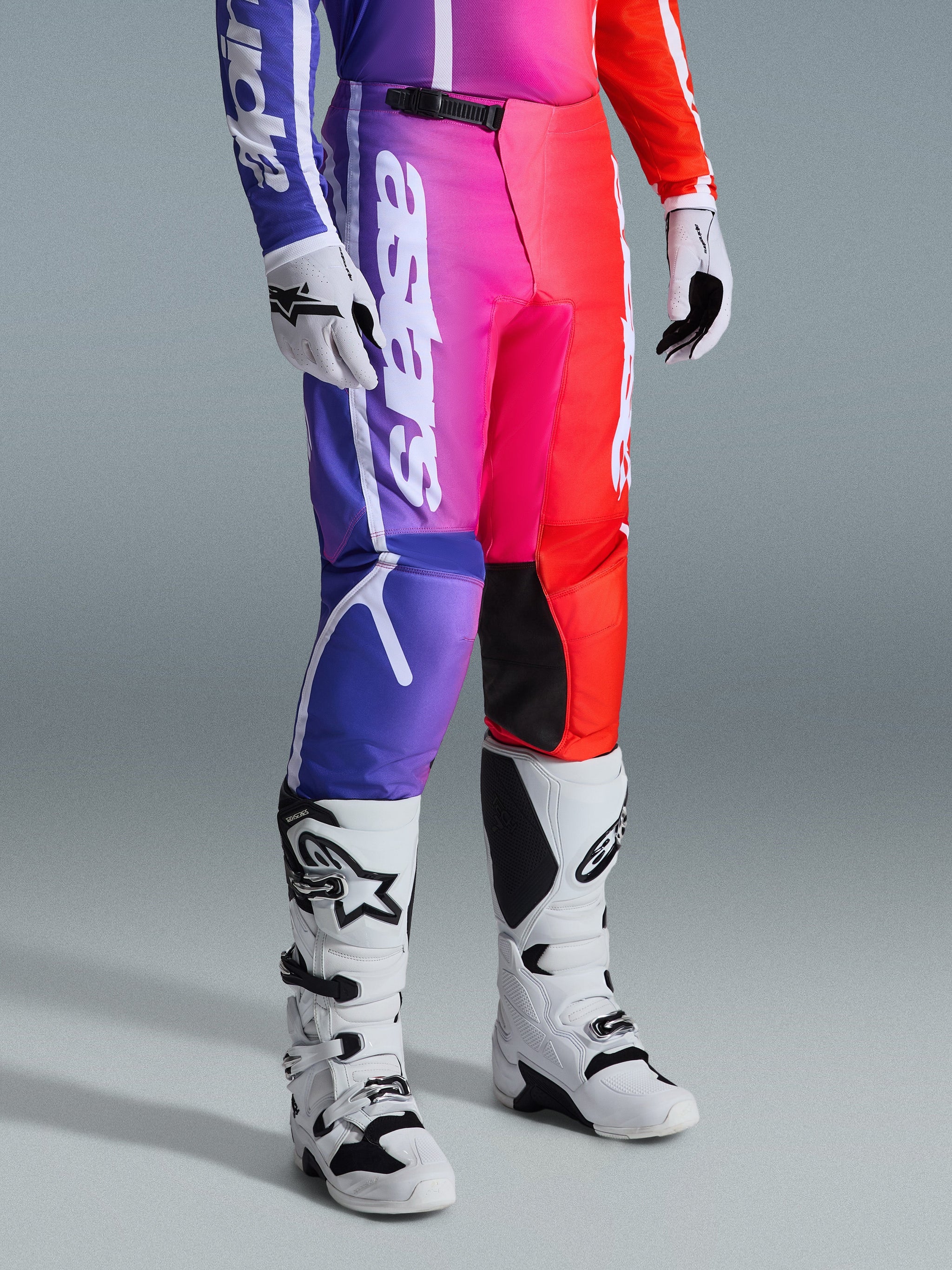 Alpinestars 2026 Fluid Apex byxor, motocrossbyxor, multifärgad lila, rosa och röd färgtoning, bärs med vita och svarta motocross-stövlar och handskar, vit vertikal logotyp och svarta knädetaljer, högpresterande terrängutrustning