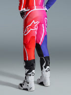 Alpinestars 2026 Fluid Apex-byxor, motocrossbyxor, flerfärgade i rött, rosa och lila, vy bakifrån av förare som bär byxor med vita och svarta motocross-stövlar, stor vit Alpinestars-logotyp, livfull tonad design