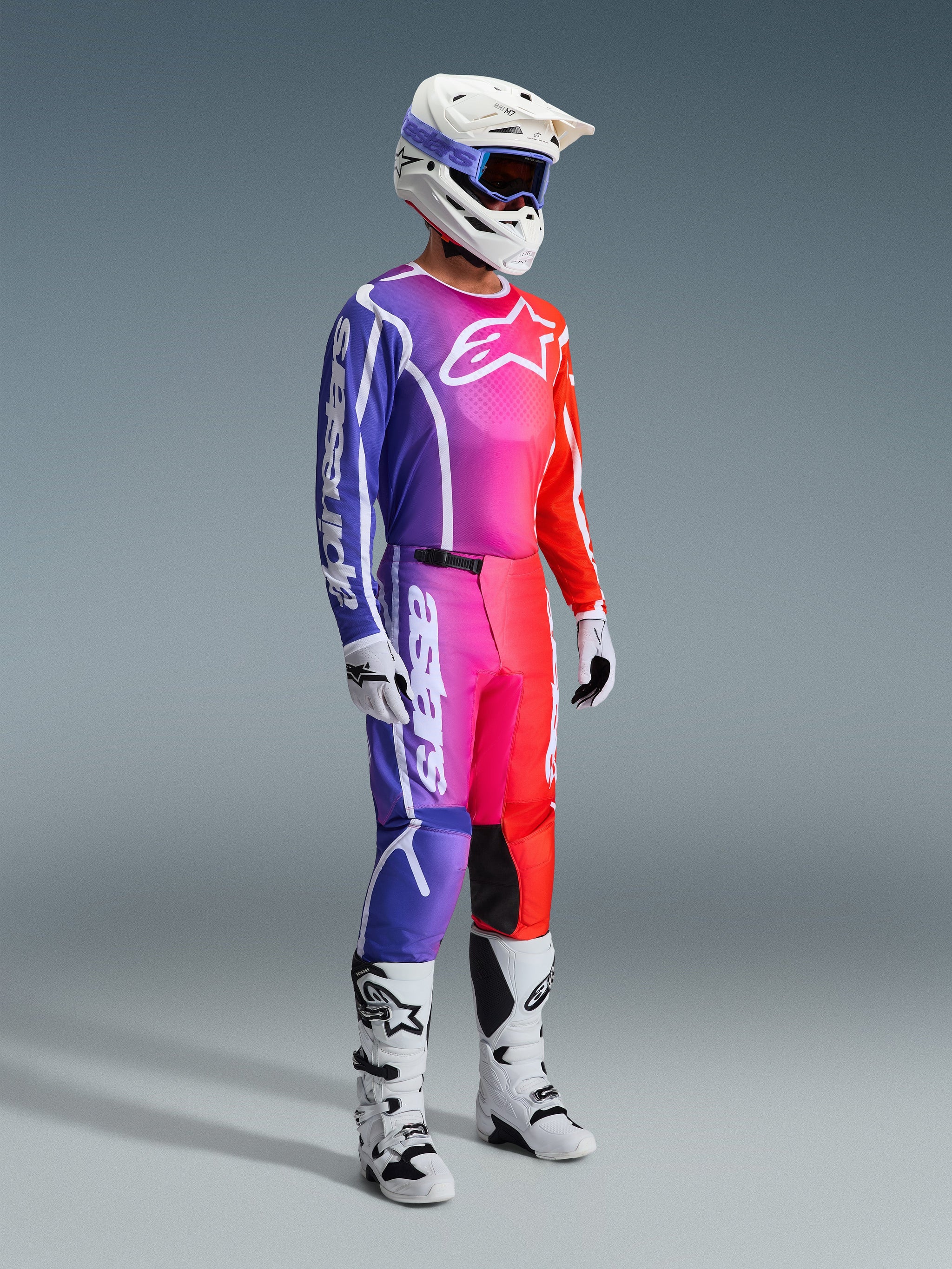 Alpinestars 2026 Fluid Apex-byxor, motocrossbyxor, flerfärgad gradient i lila, rosa och orange, bärs med matchande tröja, vit hjälm och vita stövlar, med framträdande vit logotyp och ergonomisk design för terrängkörning