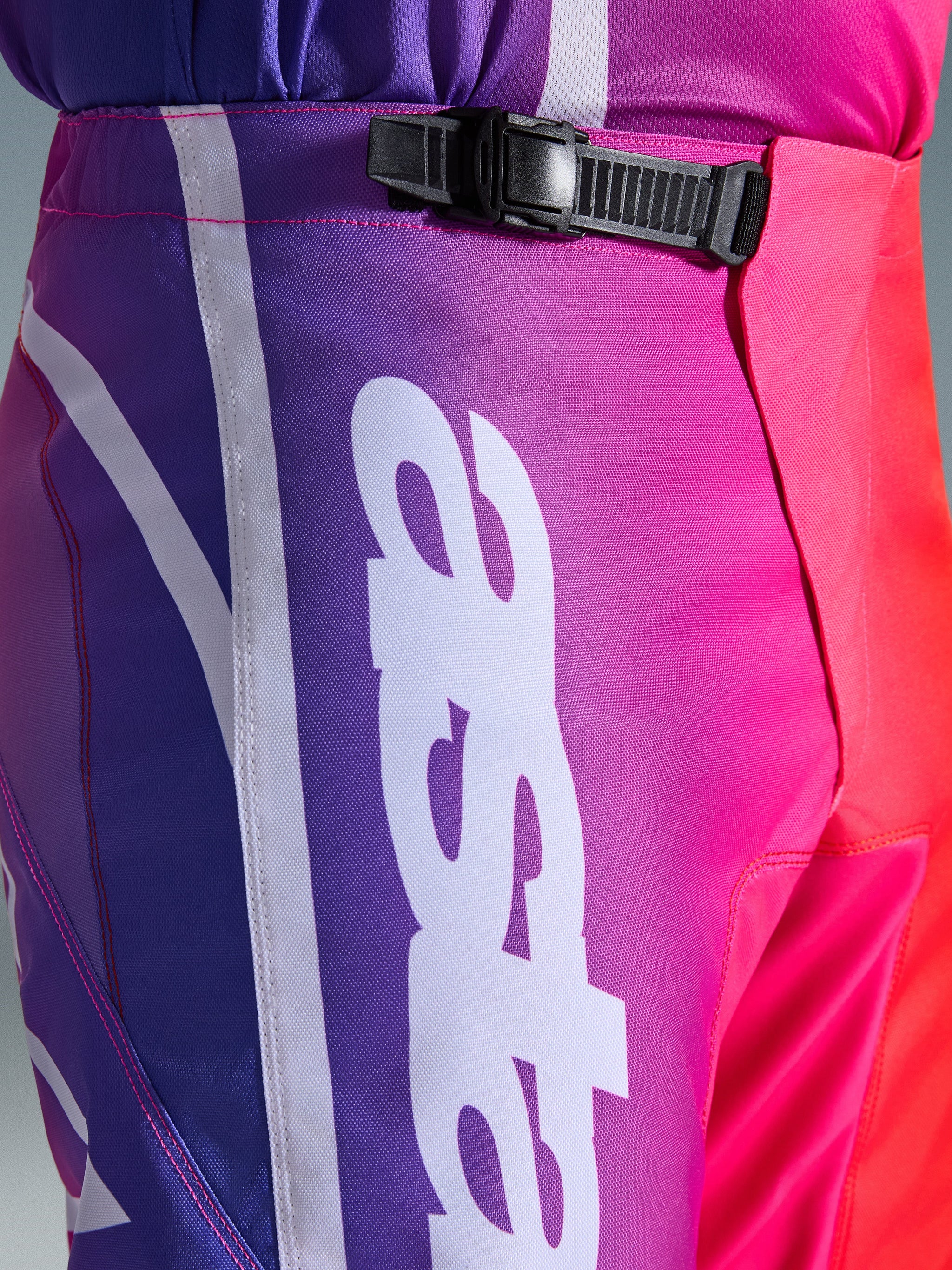 Alpinestars 2026 Fluid Apex byxor, motocrossbyxor, flerfärgade i lila, rosa och rött, närbild på midje- och höftpartiet med ett svart spänne med spärr och en stor vit vertikal Alpinestars-logotyp, livfull gradientdesign med slitstark textur i polytyg