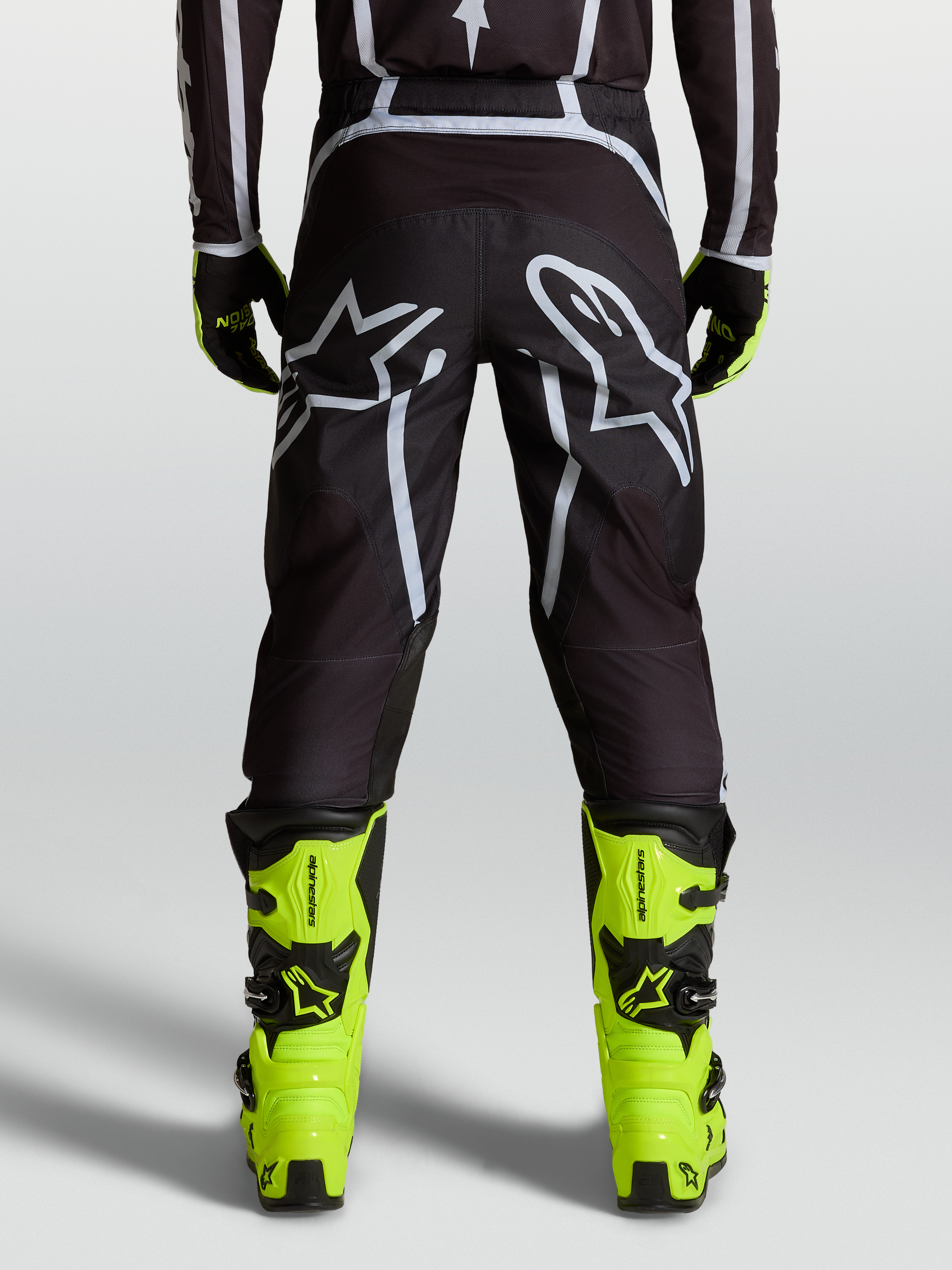 Alpinestars Fluid Apex V2 byxor, svarta och silverfärgade motocrossbyxor för herrar, sedda bakifrån på modell med neongula stövlar, ergonomisk passform och stor Astars-logotyp.