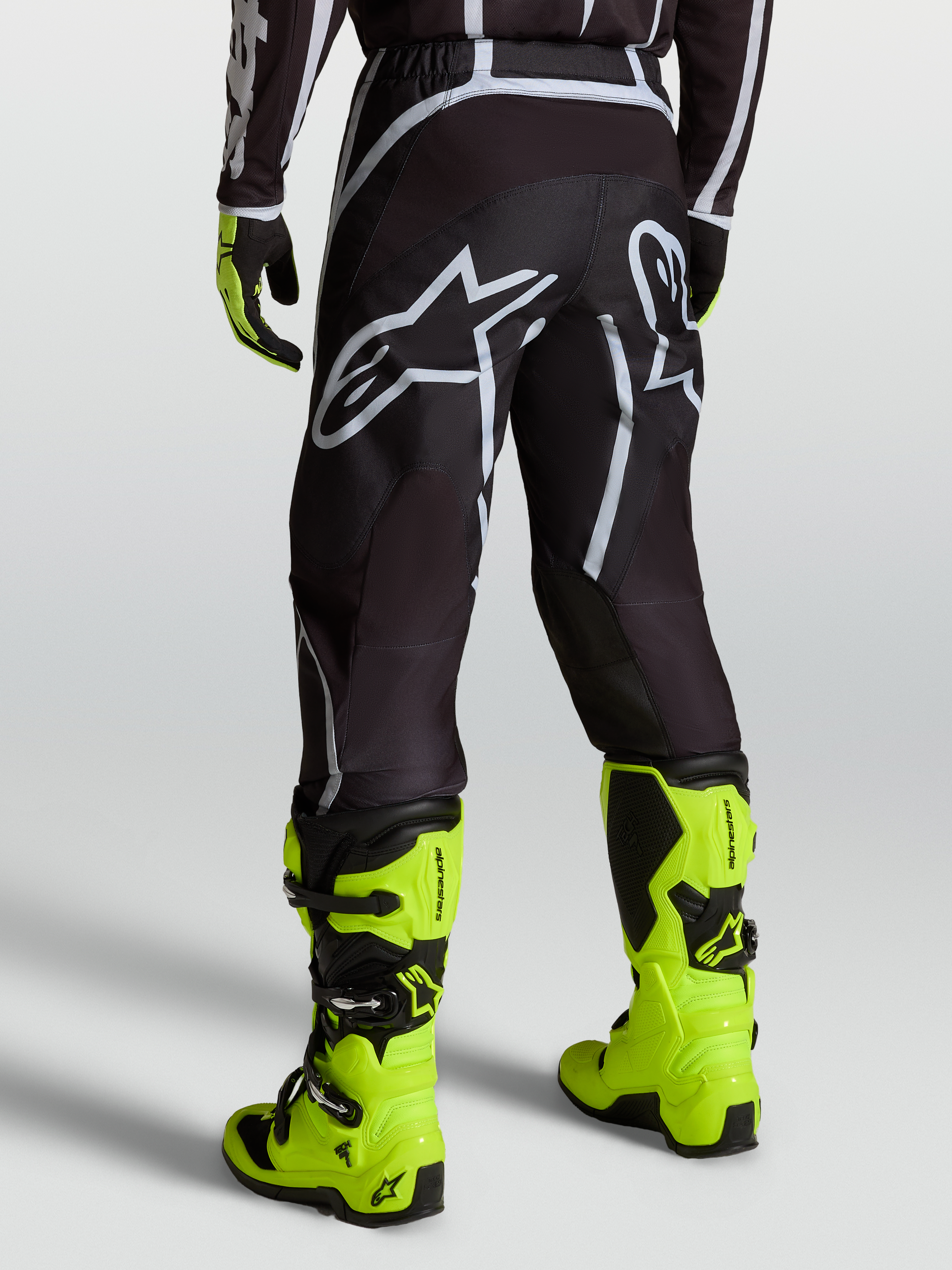 Alpinestars Fluid Apex V2 byxor, motocrossbyxor för herr i svart och silver, sedda bakifrån med neongula stövlar.