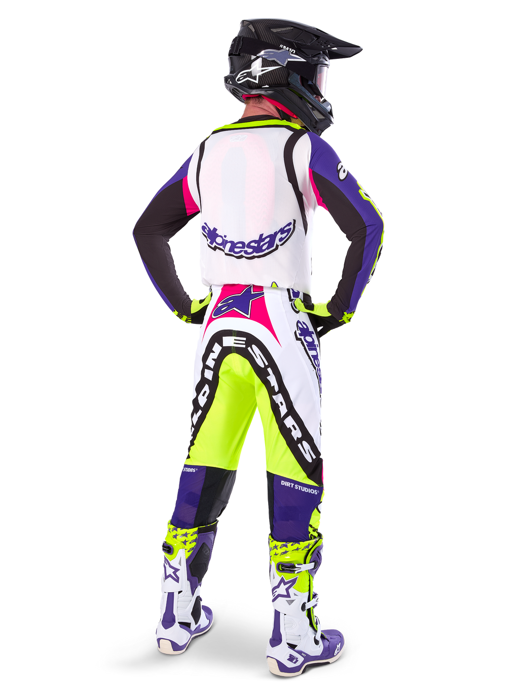 Motocrossförare i Alpinestars Limited Edition Supertech Dirt Studios® byxor i färgerna White Purple och Yellow Fluo, sedd bakifrån med matchande tröja, stövlar och svart kolfiberhjälm, professionell offroad-racingutrustning med kraftfull grafik och neonaccenter.