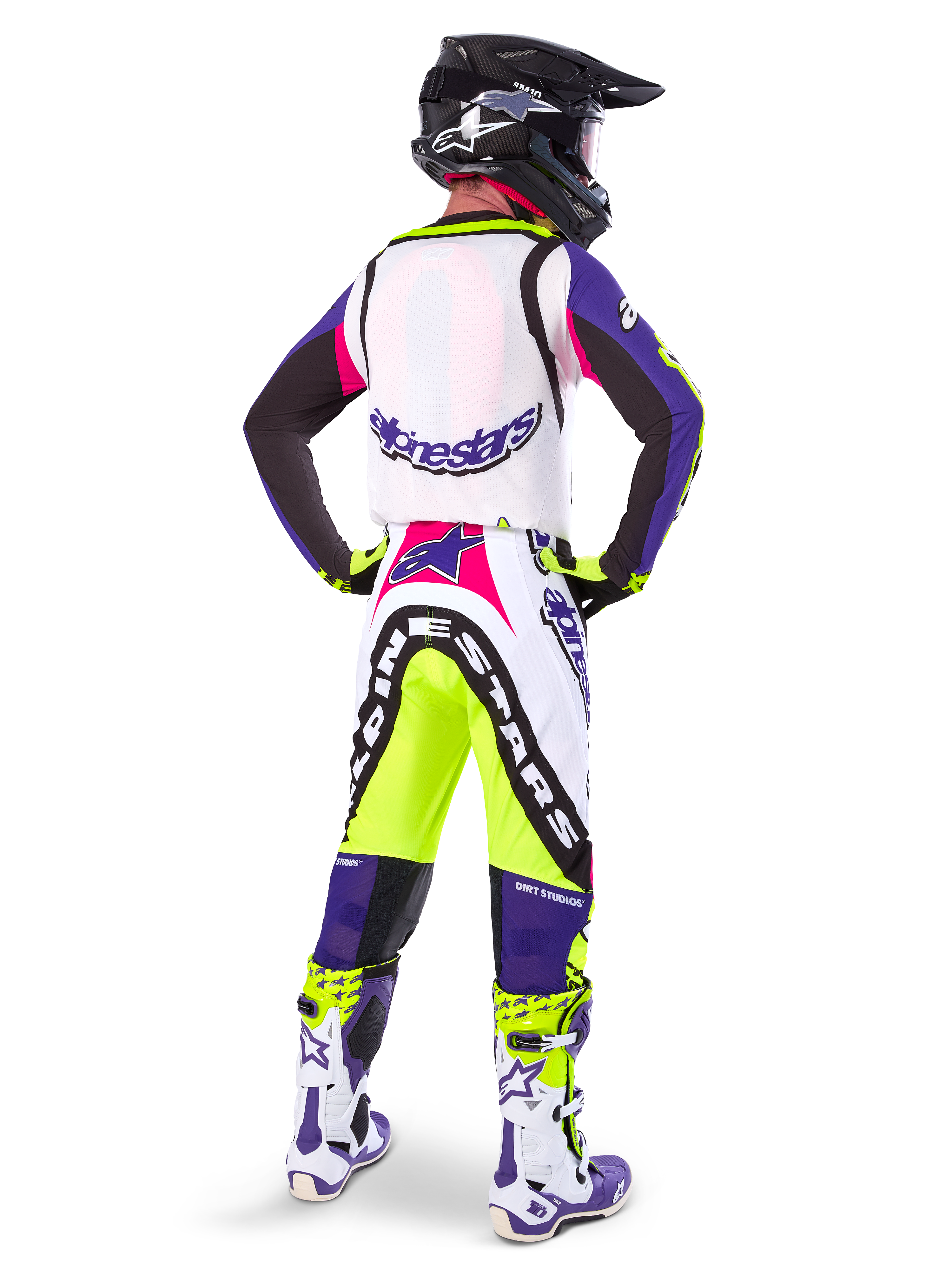 Motocrossförare i Alpinestars Limited Edition Supertech Dirt Studios® byxor i färgerna White Purple och Yellow Fluo, sedd bakifrån med matchande tröja, stövlar och svart kolfiberhjälm, professionell offroad-racingutrustning med kraftfull grafik och neonaccenter.
