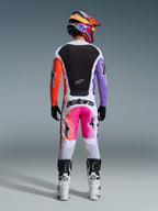 Motocrossförare i komplett racingutrustning sedd bakifrån, Alpinestars 2026 Racer Air Portl-tröja med vita, svarta, orangea och lila paneler, matchande vita byxor med rosa detaljer och 'astars'-branding, vita och svarta stövlar samt en färgstark multifärgad hjälm, koordinerad outfit för motocross.