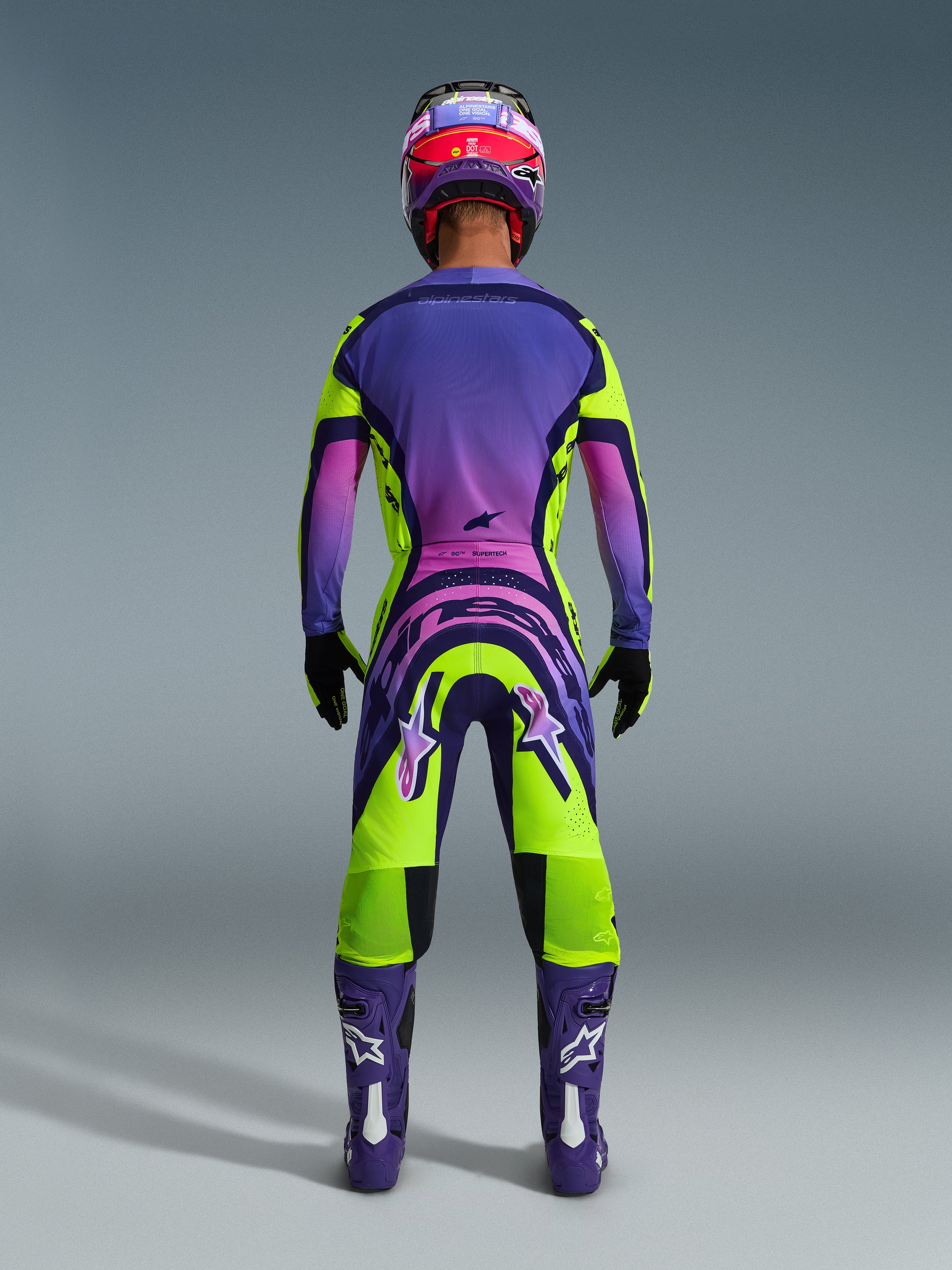 Alpinestars 2026 Supertech Vista Gear Set, back view of a motocross rider wearing a vibrant purple, pink, and lime green gradient racing outfit, featuring matching jersey and pants with bold branding, coordinated purple and pink helmet, purple boots, and black gloves.