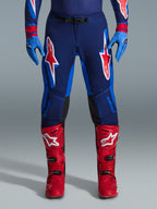 Alpinestars 2026 Supertech Vista-byxor, motocrossbyxor, mörkblå, Hot Coral och koboltblå, framifrån vy av förare i prestandautrustning med matchande blå handskar och röda motocross-stövlar, med framträdande röda och vita Alpinestars-logotyper på låren.